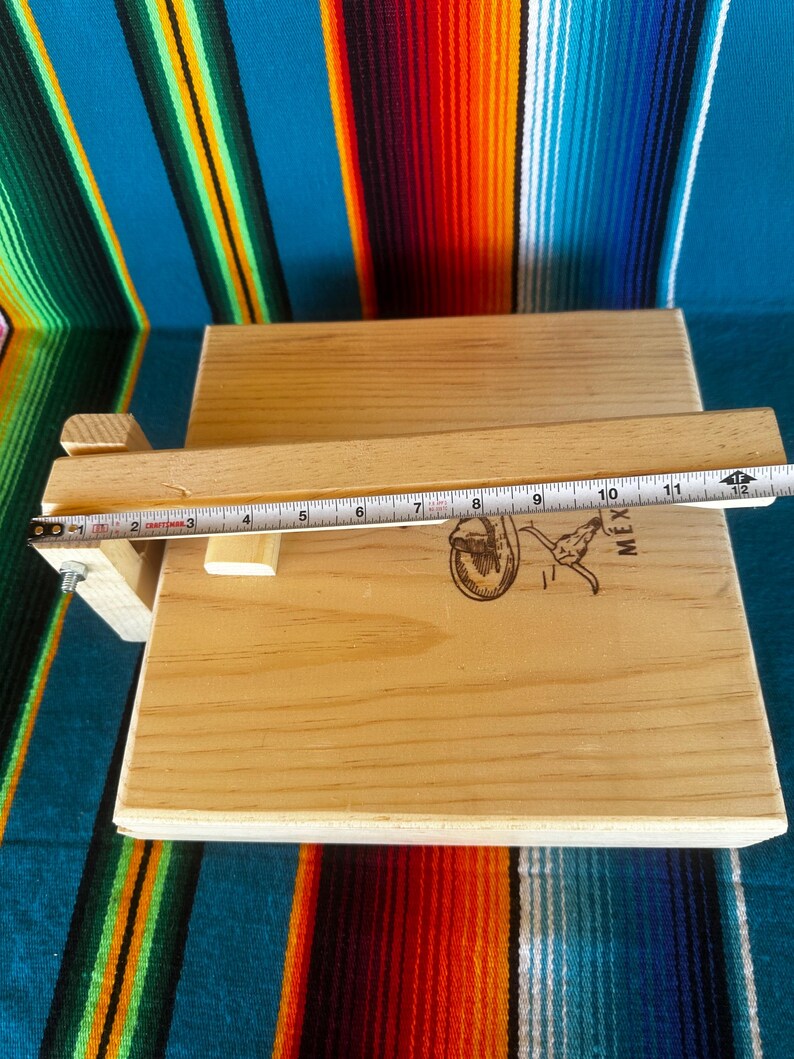 Puede incluir: Prensa de tortillas de madera clara con tapa con bisagras y asa. La prensa tiene un logotipo estampado en la superficie. Una cinta m&eacute;trica muestra una longitud de aproximadamente 30 cm.
