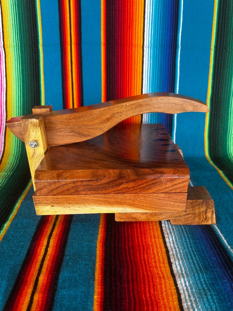 Op de afbeelding: Een houten tortillapers met een rechthoekige basis en een gebogen handvat. De pers is gemaakt van een rijk, donkerbruin hout, dat de natuurlijke nerf laat zien. De achtergrond is een kleurrijk gestreept textiel.
