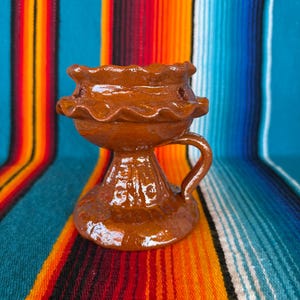 May include: A brown glazed ceramic oil lamp featuring a wide, ruffled rim, a central bowl section, a sturdy pedestal base, and a single curved handle on the side. The surface has a glossy finish with visible texture and small circular indentations.