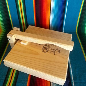 Puede incluir: Una prensa de tortillas de madera clara con una tapa con bisagras y un asa. La prensa es rectangular y presenta una imagen decorativa y la palabra "MEXI". El fondo es un textil a rayas colorido.