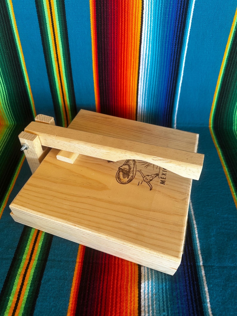 Puede incluir: Una prensa de tortillas de madera clara con una tapa con bisagras y un asa. La prensa es rectangular y presenta una imagen decorativa y la palabra "MEXI". El fondo es un textil a rayas colorido.