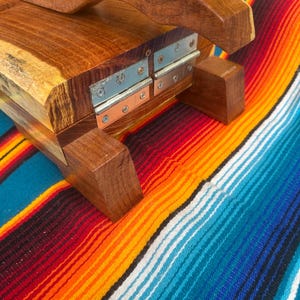 Op de afbeelding: Houten tortillapers met een gebogen handvat en metalen scharnieren. De pers is gemaakt van een rijk, donker hout en ligt op een kleurrijke gestreepte deken met rode, oranje, gele en blauwe tinten.