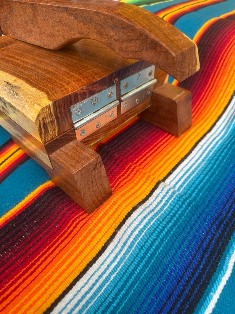Op de afbeelding: Houten tortillapers met een gebogen handvat en metalen scharnieren. De pers is gemaakt van een rijk, donker hout en ligt op een kleurrijke gestreepte deken met rode, oranje, gele en blauwe tinten.