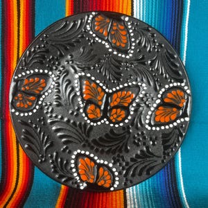 May include: A black, round decorative plate with embossed floral patterns and orange butterfly designs. The butterflies are accented with white dots. The plate is set against a colorful striped background.