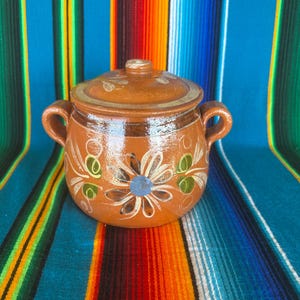May include: A small, round, brown ceramic pot with a lid. The pot features a floral design in white, green, and blue. It has two small handles and is likely used for cooking or serving. The background is a colorful striped textile.