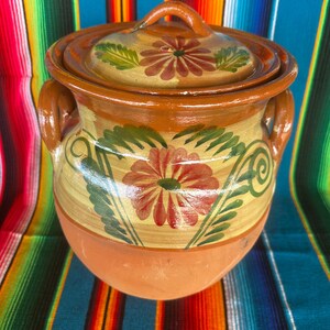 May include: A handcrafted, terracotta cooking pot with a lid. The pot features a floral design in red and green on a yellow background, with brown accents. It has two handles and a matching lid with a handle.
