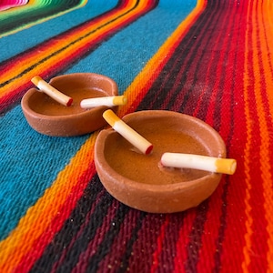 May include: Two round, terracotta-colored clay ashtrays, each featuring a small notch on the rim. Inside each ashtray rest two cylindrical, off-white objects with yellow and red tips, resembling cigarettes, placed against the inner walls.