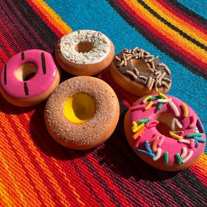 May include: An overhead shot displays five colorful donuts on a vibrant, striped textile. The donuts feature various toppings, including pink frosting with sprinkles, chocolate frosting with sprinkles, and white frosting.