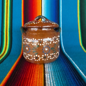 May include: A small, brown ceramic pot with a lid, adorned with white, blue, and green painted details. The pot has two small handles and a matching lid. The background is a colorful striped textile.