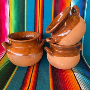 May include: Three round terracotta pots with two small handles each. The pots feature a natural matte clay base and a glossy, dark brown glazed upper section and interior. The vessels have a wide, open rim and a rounded, bulbous body shape.