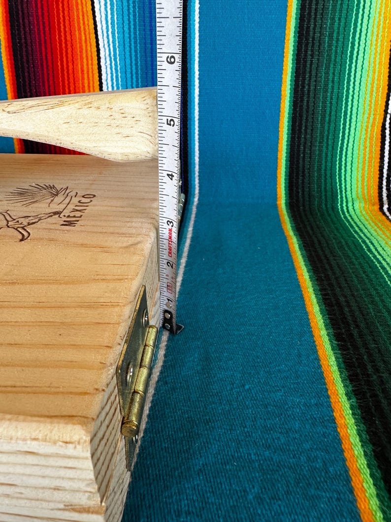 Puede incluir: Prensa para tortillas de madera clara con mango. La prensa mide aproximadamente 13 cm de alto. La palabra "MEXICO" es visible en el lateral. El fondo es un textil a rayas colorido.