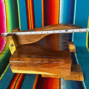 Op de afbeelding: Een handgemaakte houten tortillapers, ongeveer 35,5 cm lang. De pers is gemaakt van donkerbruin hout met een gladde, gepolijste afwerking. Het ontwerp omvat een rechthoekige basis en een gebogen bovenkant, met een scharnier voor gemakkelijk gebruik. De achtergrond is een kleurrijk gestreept textiel.