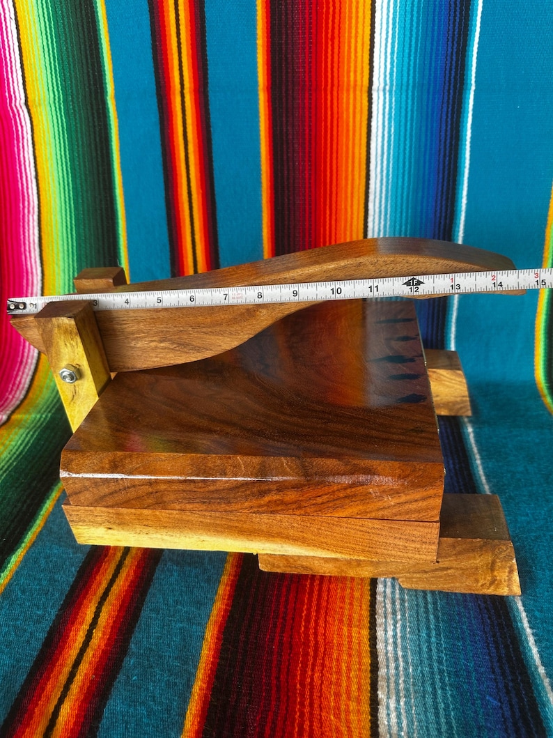 Op de afbeelding: Een handgemaakte houten tortillapers, ongeveer 35,5 cm lang. De pers is gemaakt van donkerbruin hout met een gladde, gepolijste afwerking. Het ontwerp omvat een rechthoekige basis en een gebogen bovenkant, met een scharnier voor gemakkelijk gebruik. De achtergrond is een kleurrijk gestreept textiel.
