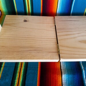 Puede incluir: Una mesa de manualidades plegable de madera con un acabado de madera clara. La mesa est&aacute; articulada en el medio y tiene una peque&ntilde;a pieza de soporte de madera adjunta al costado. La mesa se muestra sobre un textil a rayas de colores.
