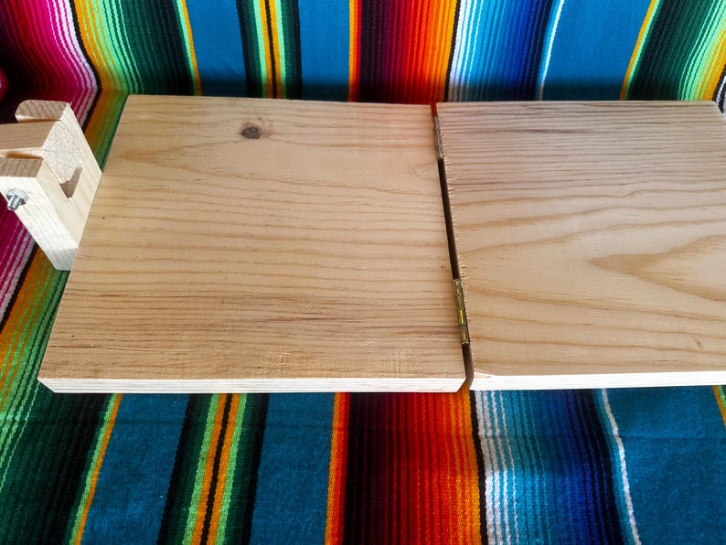 Puede incluir: Una mesa de manualidades plegable de madera con un acabado de madera clara. La mesa est&aacute; articulada en el medio y tiene una peque&ntilde;a pieza de soporte de madera adjunta al costado. La mesa se muestra sobre un textil a rayas de colores.