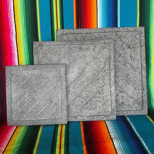 May include: Three gray, square stone tiles of varying sizes are arranged in a row. Each tile has a textured surface and a diagonal line pattern. The tiles are displayed against a colorful striped background.