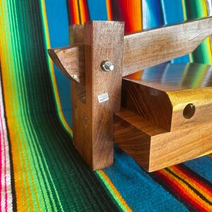 Op de afbeelding: Een houten tortillapers met een rechthoekige basis en een scharnierende bovenkant. De pers is gemaakt van donkerbruin hout en heeft een metalen bout. De achtergrond is een kleurrijk gestreept textiel.