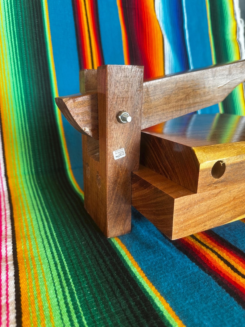 Op de afbeelding: Een houten tortillapers met een rechthoekige basis en een scharnierende bovenkant. De pers is gemaakt van donkerbruin hout en heeft een metalen bout. De achtergrond is een kleurrijk gestreept textiel.