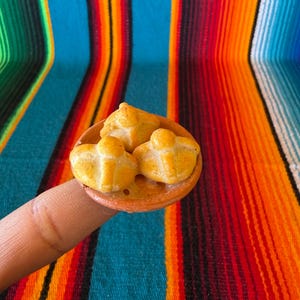 May include: A miniature ring featuring a small, round, terracotta-colored plate topped with three golden-brown, rounded bread rolls. Each roll has a distinct cross-shaped dough decoration on top, mimicking traditional pan de muerto bread.