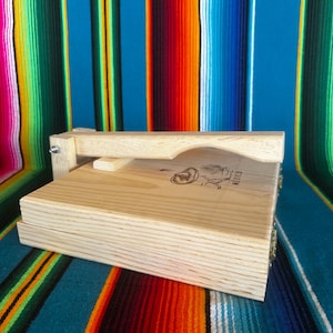 Puede incluir: Una prensa de tortillas de madera clara con un asa. La prensa es rectangular con una tapa con bisagras y un dise&ntilde;o grabado en la parte superior. El fondo es un textil a rayas de colores.