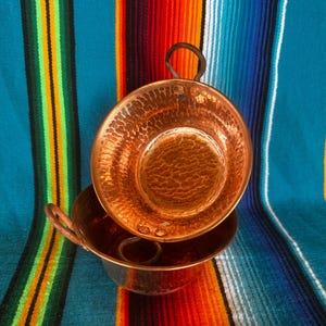 May include: Two hammered copper bowls with handles, one resting inside the other. The bowls have a textured surface and a warm, reddish-brown color. The background is a colorful striped textile in shades of blue, orange, and green.