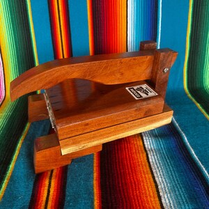 Op de afbeelding: Een houten tortillapers met een handvat en een rechthoekige basis. De pers is gemaakt van lichtbruin hout en heeft een label met "Mexico". De pers staat tegen een kleurrijke gestreepte achtergrond.