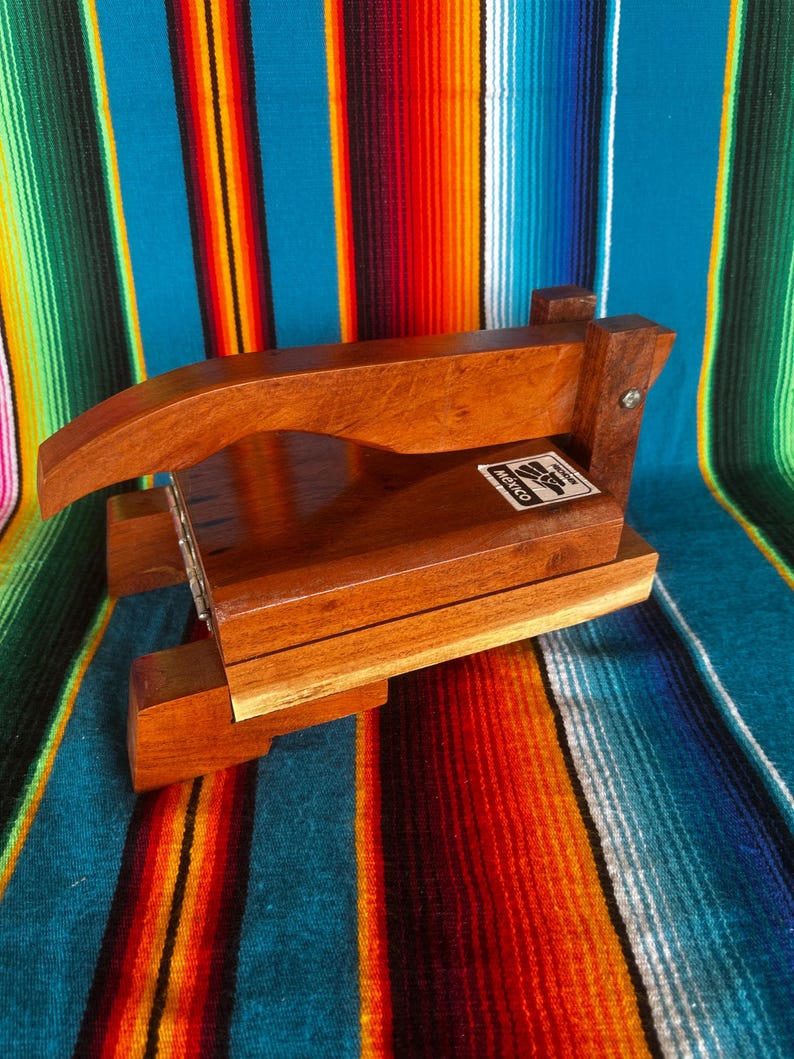 Op de afbeelding: Een houten tortillapers met een handvat en een rechthoekige basis. De pers is gemaakt van lichtbruin hout en heeft een label met "Mexico". De pers staat tegen een kleurrijke gestreepte achtergrond.