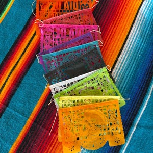 May include: A stack of rectangular paper banners featuring intricate cut-out designs of skulls, flowers, and the text DIA DE LOS MUERTOS. The banners are in vibrant shades of orange, pink, purple, blue, black, white, lime green, and yellow.