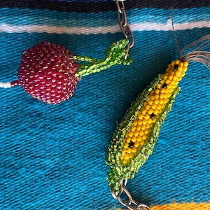 May include: Two beaded keychains: a red cherry with green leaves and a yellow corn on the cob with green husks. The keychains are made of small beads and are attached to silver-colored chains. The background is a blue and turquoise woven fabric.