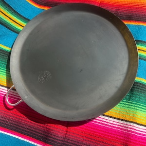 May include: A dark gray, round griddle pan with a single, curved handle. The pan has a slightly textured surface and a small logo near the handle. The pan is sitting on a colorful, striped textile.