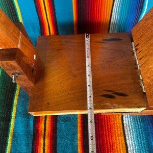 Op de afbeelding: Een open houten kist met een scharnierend deksel, met een donkerhouten interieur. Een metalen meetlint geeft aan dat het interieur ongeveer 28 cm lang is. De kist staat tegen een kleurrijke gestreepte textielachtergrond.