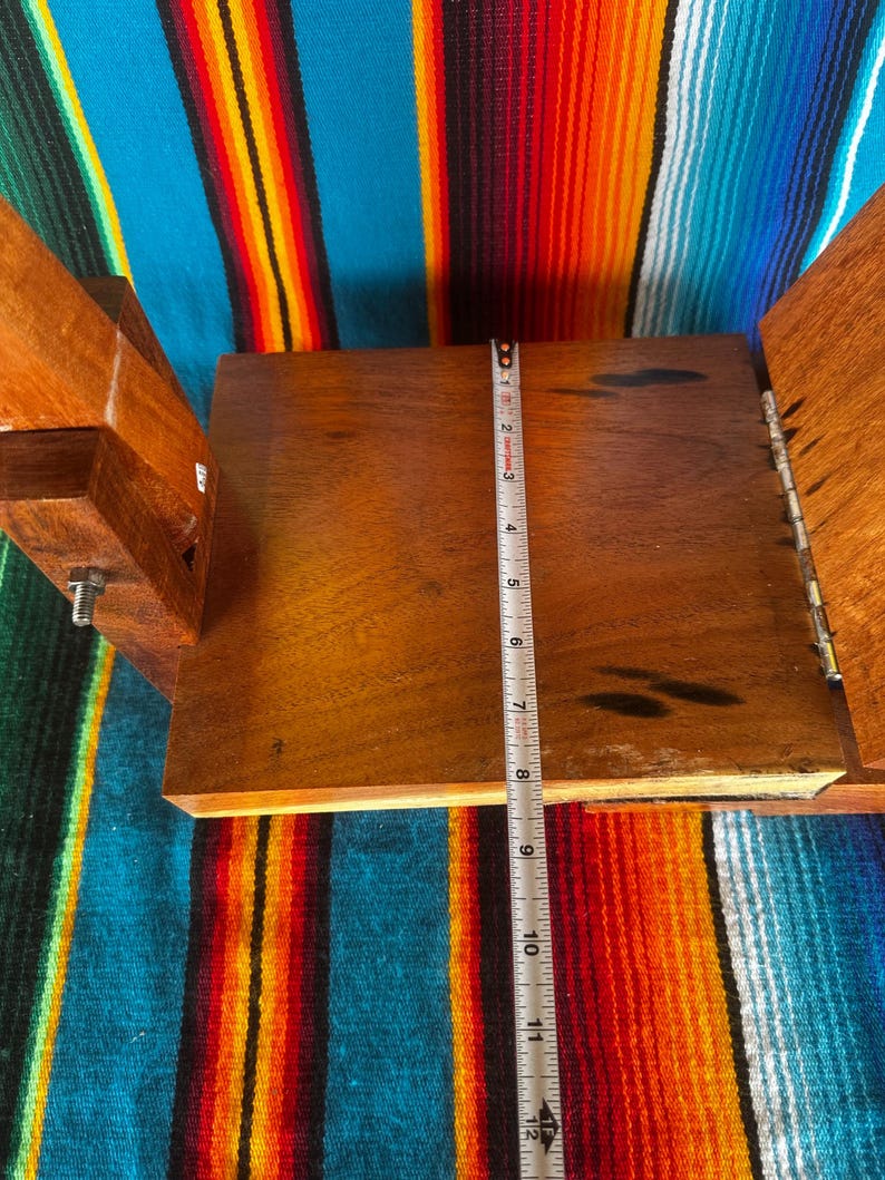 Op de afbeelding: Een open houten kist met een scharnierend deksel, met een donkerhouten interieur. Een metalen meetlint geeft aan dat het interieur ongeveer 28 cm lang is. De kist staat tegen een kleurrijke gestreepte textielachtergrond.
