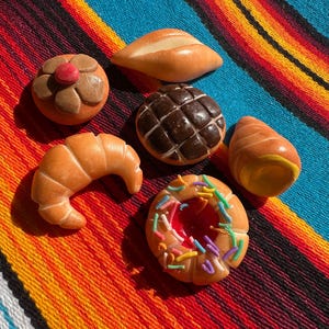 May include: A collection of miniature ceramic baked goods, including a croissant, a donut with sprinkles, a bread roll, a concha, a bolillo, and a bread roll. The items are arranged on a colorful striped textile.