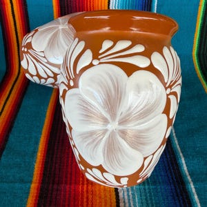 May include: Two terracotta-colored ceramic vases featuring a hand-painted white floral pattern. The vases have a rounded body and a flared rim, with the white petals covering the surface in a repeating design against the reddish-brown clay base.