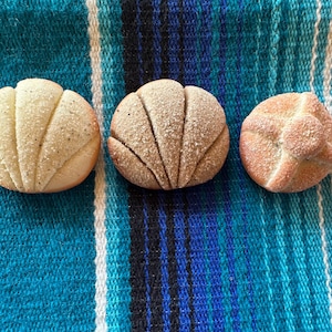 May include: Three Mexican concha pastries, each with a unique color and texture, rest on a blue, white, and yellow striped textile. The conchas are round, with a shell-like pattern on top, and are dusted with sugar.