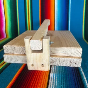 Puede incluir: Una prensa de tortillas de madera con un mango y un dise&ntilde;o de bisagra. La prensa est&aacute; hecha de madera de color claro y est&aacute; dise&ntilde;ada para aplanar la masa en tortillas. La prensa se muestra sobre un fondo a rayas colorido.