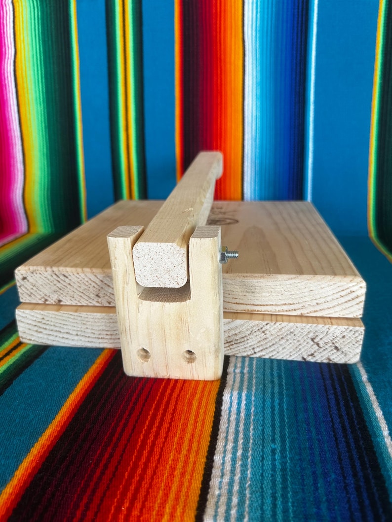 Puede incluir: Una prensa de tortillas de madera con un mango y un dise&ntilde;o de bisagra. La prensa est&aacute; hecha de madera de color claro y est&aacute; dise&ntilde;ada para aplanar la masa en tortillas. La prensa se muestra sobre un fondo a rayas colorido.