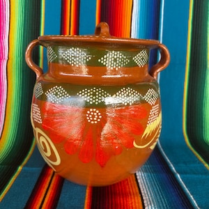 May include: A hand-painted, terracotta cooking pot with a lid. The pot features a brown base, green and white dotted bands, and a vibrant red floral design. It has two side handles and a top handle.