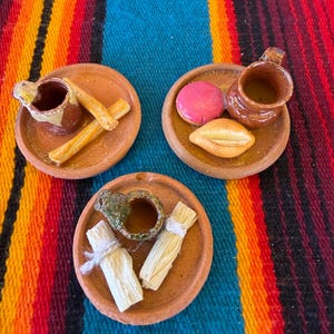 May include: Three miniature terracotta plates, each holding a small ceramic mug and food items. One plate features two churros, another holds a pink round pastry and a bread roll, and the third displays two small tamales tied with white string.