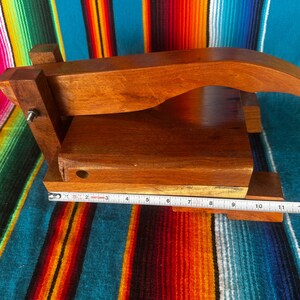 Op de afbeelding: Een houten tortillapers met een natuurlijke afwerking. De pers is rechthoekig met een gebogen handvat en meet ongeveer 28 cm lang. De pers staat tegen een kleurrijke gestreepte achtergrond.