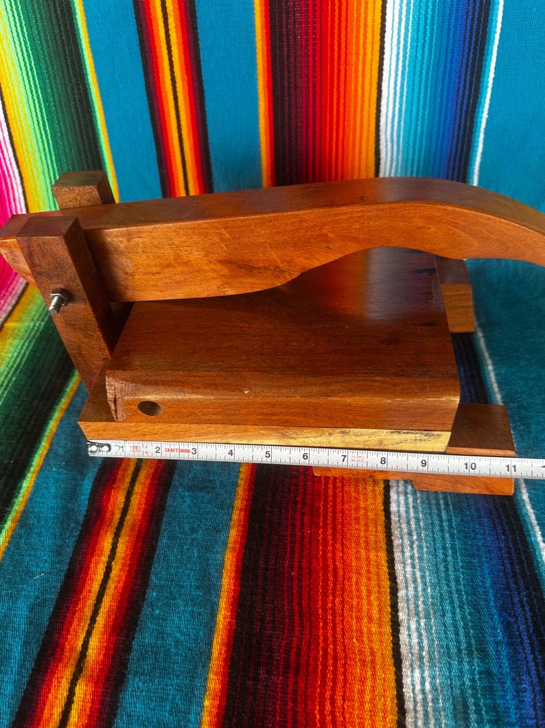 Op de afbeelding: Een houten tortillapers met een natuurlijke afwerking. De pers is rechthoekig met een gebogen handvat en meet ongeveer 28 cm lang. De pers staat tegen een kleurrijke gestreepte achtergrond.