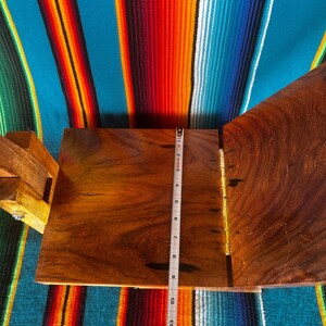 Op de afbeelding: Een houten kist met een scharnierend deksel, open om de binnenkant te onthullen. Een meetlint toont dat de kist ongeveer 25 cm lang is. De kist is gemaakt van donker hout en heeft messing scharnieren. De achtergrond is een kleurrijk gestreept textiel.