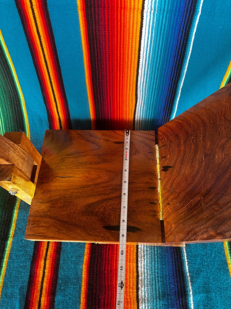 Op de afbeelding: Een houten kist met een scharnierend deksel, open om de binnenkant te onthullen. Een meetlint toont dat de kist ongeveer 25 cm lang is. De kist is gemaakt van donker hout en heeft messing scharnieren. De achtergrond is een kleurrijk gestreept textiel.