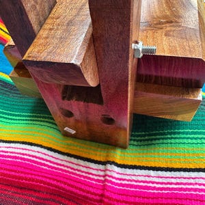 Op de afbeelding: Een handgemaakt houten object met een rijke, donkerbruine afwerking. De structuur is geassembleerd met in elkaar grijpende stukken en een metalen bout. Het object rust op een kleurrijk gestreept textiel, dat het ontwerp en het vakmanschap laat zien.