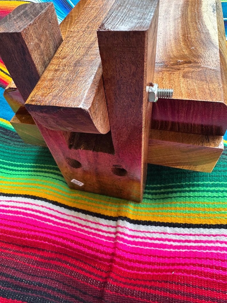 Op de afbeelding: Een handgemaakt houten object met een rijke, donkerbruine afwerking. De structuur is geassembleerd met in elkaar grijpende stukken en een metalen bout. Het object rust op een kleurrijk gestreept textiel, dat het ontwerp en het vakmanschap laat zien.