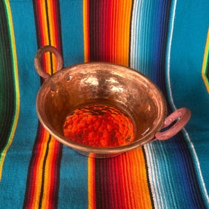 May include: A hammered copper bowl with two loop handles, reflecting a vibrant orange object inside. The bowl sits on a colorful striped textile with blue, orange, and green hues, creating a striking contrast.
