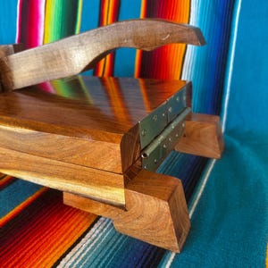 Op de afbeelding: Een houten tortillapers met een scharnierend ontwerp en een gebogen handvat. De pers is gemaakt van gepolijst, donkergebeitst hout en heeft metalen scharnieren. De achtergrond is een kleurrijk gestreept textiel.