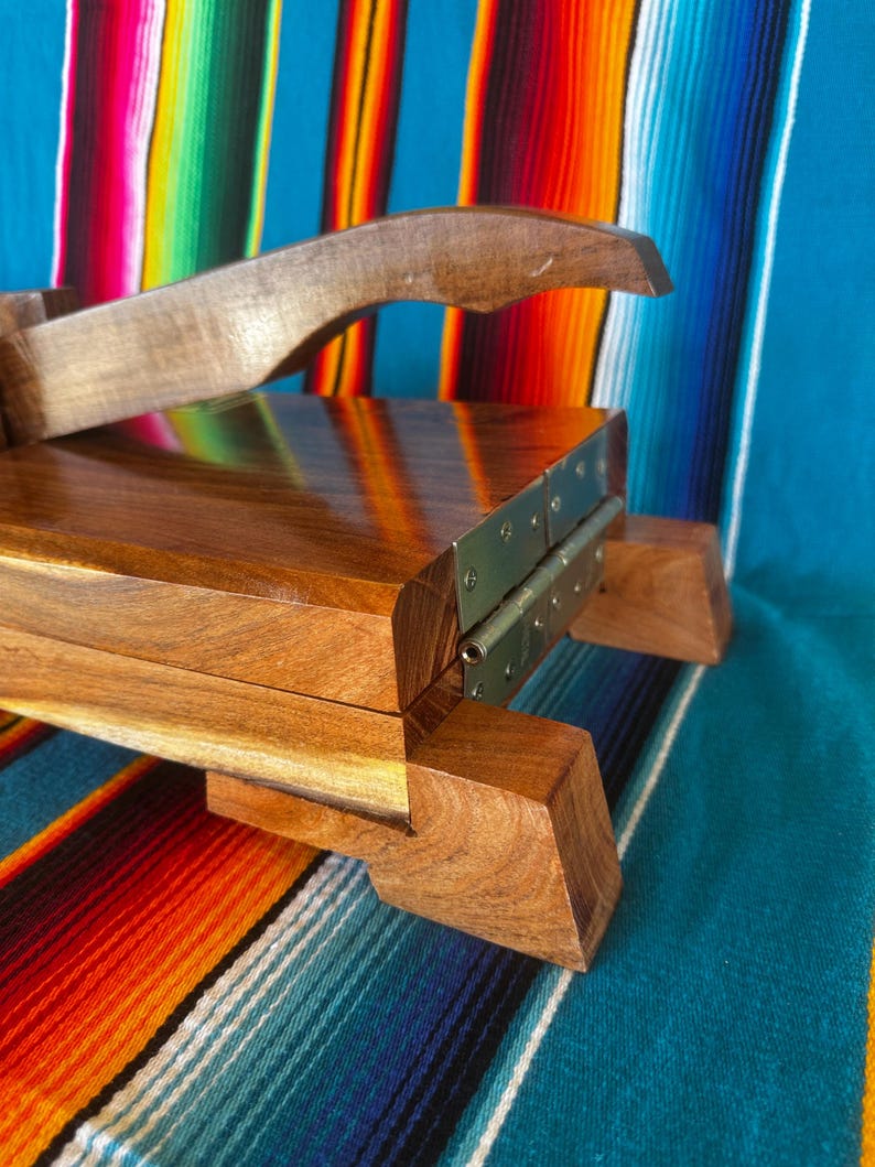 Op de afbeelding: Een houten tortillapers met een scharnierend ontwerp en een gebogen handvat. De pers is gemaakt van gepolijst, donkergebeitst hout en heeft metalen scharnieren. De achtergrond is een kleurrijk gestreept textiel.