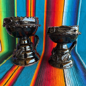 May include: Two glossy black ceramic vessels with handles, each featuring a wide bowl-shaped top and a tapered base. The exterior surfaces are decorated with raised leaf and floral motifs, and the rims have a scalloped, textured edge design.