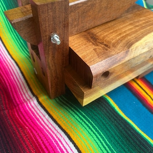 Op de afbeelding: Een houten tortillapers met een donkerbruine afwerking. De pers heeft een rechthoekige basis en een scharnierend bovendeel met een handvat. Een metalen bout zet de componenten van de pers vast. De pers staat tegen een kleurrijke gestreepte textielachtergrond.